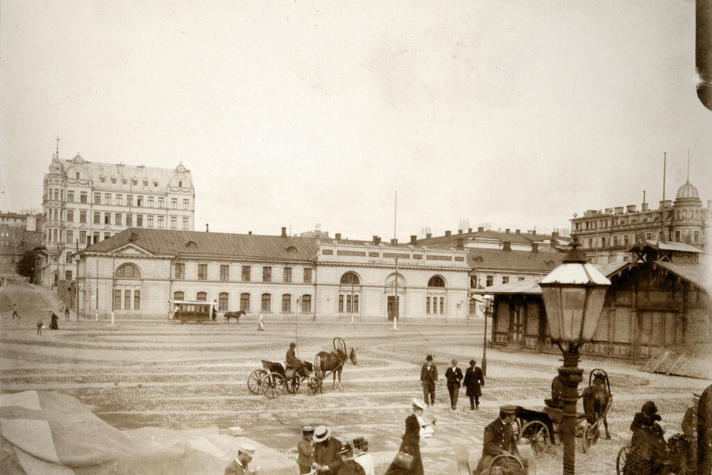 Hevosajureita rattaineen ja herrasväkeä Eteläsatamassa Pakkahuoneentorilla. Etualalla kaasulla toimiva katulyhty. Kuvaaja: Helsingin kaupunginmuseo / Alexander Eugén Maconi