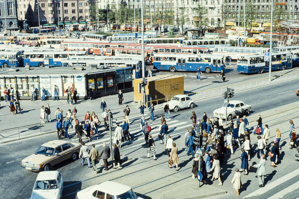 Fotgängare på väg över Brunnsgatan längs skyddsvägen, i bakgrunden Järnvägstorgets busshållplatser och busstrafik. I bildens högra övre hörn syns en rörkonstruktion som hör till metrotunnelns sprängningsarbeten som utfördes 1976–1977 i Gloets krosszon. Foto: Helsingfors stadsmuseum / Jalmari Aarnio