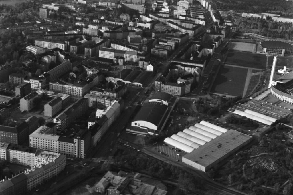 Mannerheimvägen. Sned flygbild från Tölöviken mot nordost. I förgrunden syns sockerbrukets tomt med en byggnad i kanten mot Helsingegatan. I mitten Mässhallen, till höger på Tallbackafältet den tillfälliga Mässhallen C, dvs. Folkens hall. I bakgrunden i mitten syns Mannerheimvägen. Bilden är från år 1970. Foto: Helsingfors stadsmuseum / Simo Rista