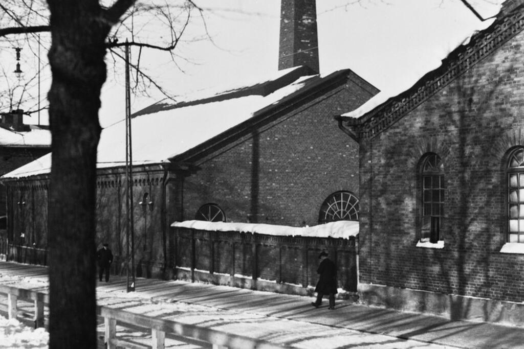 Fotgängare vid gasverket. Bilden är tagen från Henriksparken som låg mellan Västra och Östra Henriksgatan, alltså i mitten av nuvarande Mannerheimvägen.  Foto: Helsingfors stadsmuseum / R. Roos