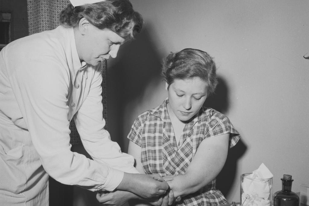 Valkohilkkainen terveydenhoitaja pistämässä rokotetta naisen käsivarteen.  Pöydällä naisen vieressä rokotustarpeita.