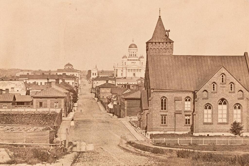 Helsinki oli 1870-luvulla puutalovaltainen kaupunki, jonka maisemassa erottuivat kirkot ja julkisen vallan kivitalot. Kuvan oikeassa kulmassa on vuonna 1864 valmistunut Saksalainen kirkko. Vasemmalla näkyy Svenska Normallyceumin halkopinoja.  Kuvaaja: Helsingin kaupunginmuseo / Carl Eugen Hoffers