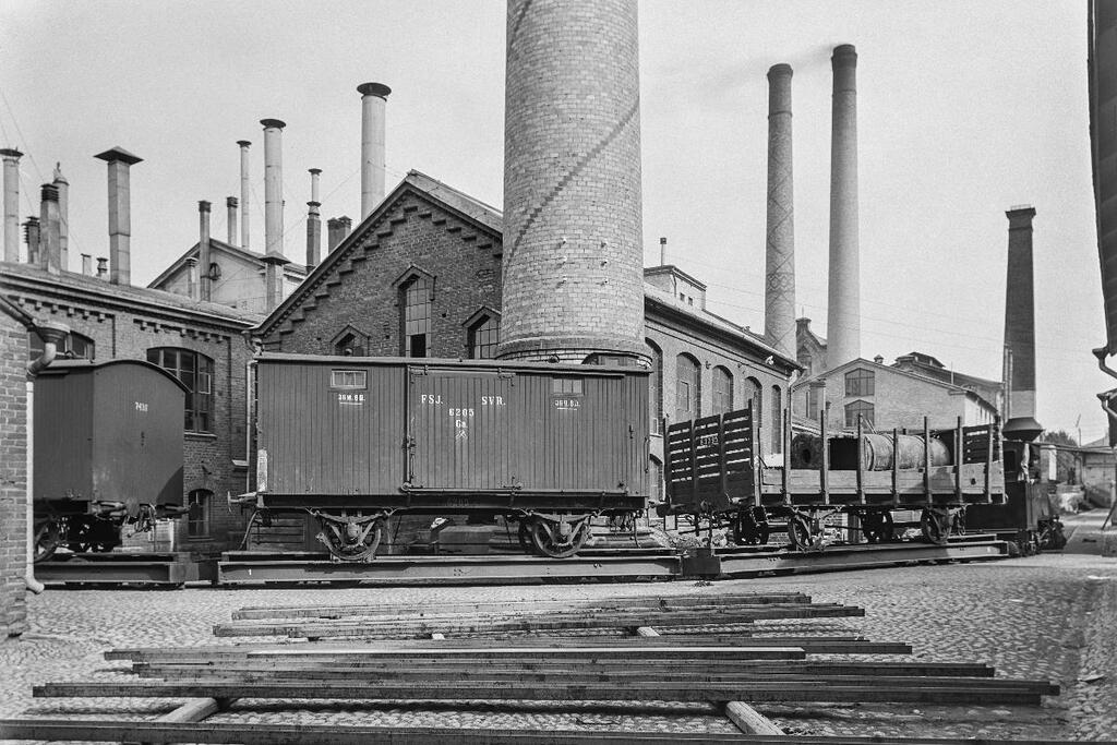 Etualalla maassa lautoja, kuvan keskellä junan tavaravaunuja ja niiden takana punatiilisiä tehdasrakennuksia savupiippuineen.