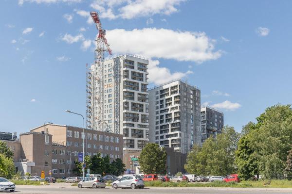 . I förgrunden syns parkerade bilar och träd. I bakgrunden reser sig tre höga våningshus som har byggställningar. Bredvid den högsta byggnaden står en lyftkran som är högre än huset.