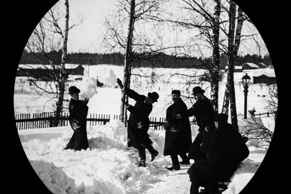 Hattuihin ja pitkiin takkeihin pukeutuvat miehet leikkivät lumisotaa pihatiellä. Taustalla näkyy koivuja, piha-aita ja kaasulyhty.