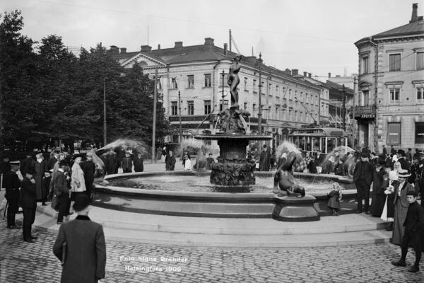 Människor har samlats kring skulpturen som står i mitten. I bakgrunden till vänster syns Esplanadparken, i mitten och till höger byggnader på Norra Esplanaden 19 och Unionsgatan 28.