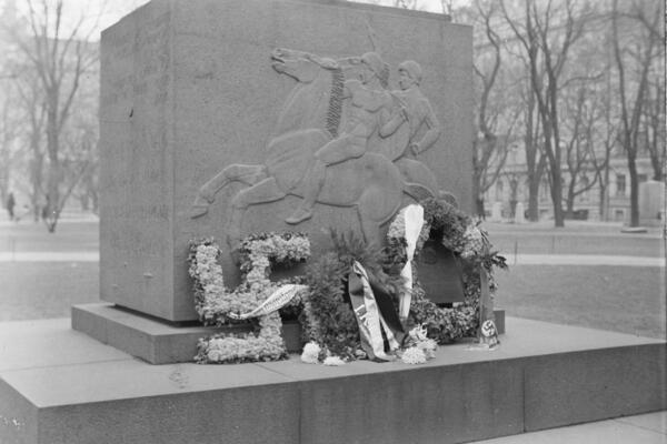 En fyrkantig minnessten med en relief som avbildar två män ridande på en häst. Framför minnesmärket har man placerat blomkransar.