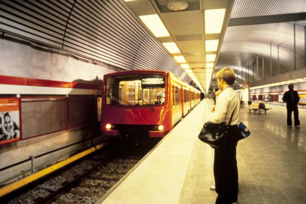 Metrojuna on juuri pysähtymässä laiturille Hakaniemessä. Etualalla metroa odottaa kahta laukkua kantava, villapaitaan pukeutunut nuori poika.