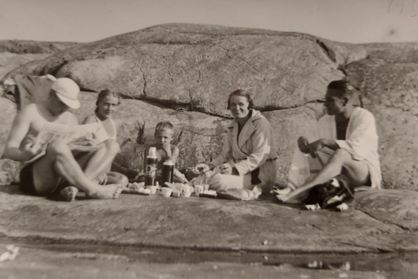 Två män och kvinnor med en pojke i mitten sitter på strandklippor med matsäck. Den ena mannen bär keps och läser en tidning. 