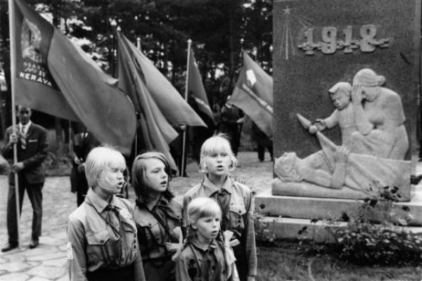Neljä nuorta pioneeriasuista tyttöä laulaa Santahaminan veljeshaudalla punaisten muistomerkin edustalla. Taustalla lippuja piteleviä miehiä.