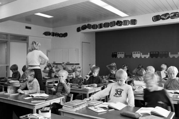 Alakoululaisia oppitunnilla luokassaan. Oppilaiden pöydillä on kirjapinoja, opettaja kiertää luokassa.