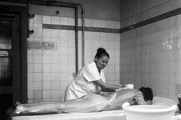 A washerwoman dressed in a white coat soaping up the back of a man lying face down. There are white tiles and a sign with the text “Using soap in the sauna or going to the sauna covered in soap is strictly prohibited” in the background.