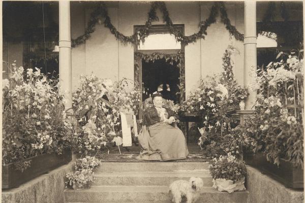 Aurora Karamzin fotograferad på sin 90-årsdag sittande framför ytterdörren till sin villa, omringad av blomplanteringar. En liten ljus hund sitter framför henne på trappan. 