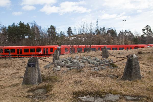 Pronssikautinen hautaröykkiö. Haudan muodostaa muutaman neliömetrin alueen kattava kasa jalkapallon kokoisia kiviä, jotka on eristetty kettingillä. Taustalla kulkee metrojuna.