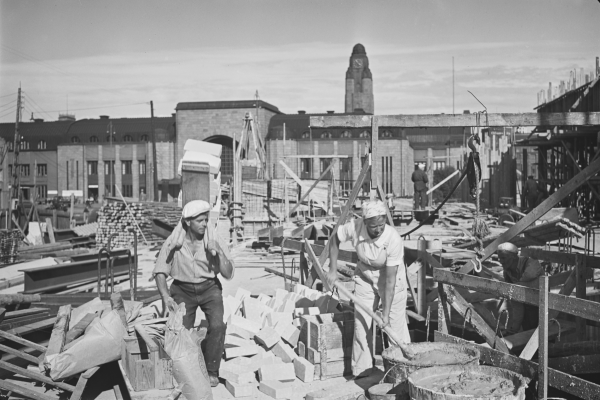 En kvinna blandar murbruk i en stor hink på byggplatsen för Sokos varuhus. I bakgrunden Järnvägsstationens byggnad.