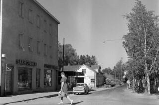 Tapanila, Veljestentie 17–21. Vasemmalla on kolmikerroksinen rapattu talo, jonka pohjakerroksessa on liikehuoneistoja. Seinässä on valomainokset SÄÄSTöPANKKI ja APTEEKKI APOTEK. Mekkoon pukeutunut nainen kävelee tien yli kohti rakennusta. Taustalla henkilöauto ja vaalea harjakattoinen puutalo, oikealla koivuja.