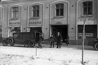 Luminen katu ja jalkakäytävä, jolle on pysähtynyt ambulanssi tai ruumiinkuljetusauto. Kolme miestä nostaa paareilla makaavaa, tummalla huovalla peitettyä ruumista auton takaosaan. Taustalla kävelee ohikulkija.