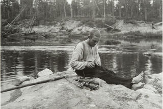 Mies istuu kalliolla veden ääressä ja avaa veitsellä raakkua. Hänen vieressään on kasa raakkuja.