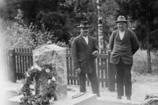 Kaksi miestä seisoo seppeleellä koristellun hautakiven vierellä.