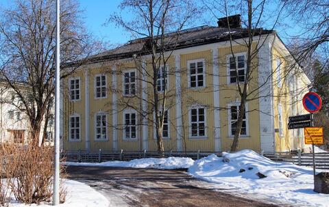 Kaksikerroksinen aumakattoinen keltaiseksi maalattu puurakennus, jossa on kahdeksanruutuiset ikkunat.