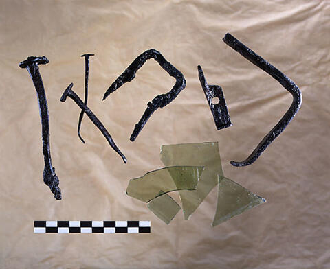 Gråaktiga glasbitar och rostiga, vridna metallföremål. Två av dem kan identifieras som spikar.