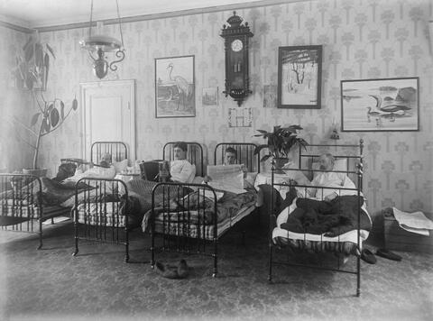 Fyra män i järnsängar bredvid varandra. En spelar flöjt, en spelar dragspel och två läser tidningar.