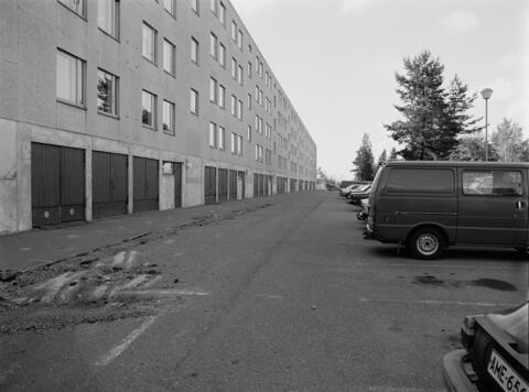 Vasemmalla on pitkä tasakattoinen elementtikerrostalo, jonka pohjakerroksessa on autotallinovia. Asuinkerroksia on neljä. Oikealla on pysäköityjä autoja.