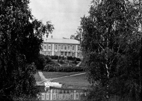 Kuvan keskellä on kaksikerroksinen kartanorakennus, joka on kuvattu vastarannalta. Talo heijastuu joen pintaan. Kuvan vasemmassa ja oikeassa reunassa on koivuja. 
