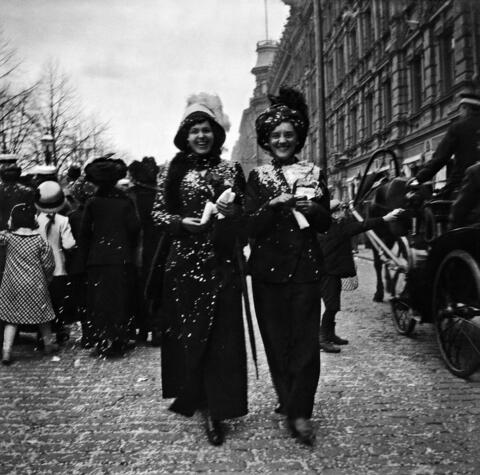 Two broadly smiling women walk along Esplanadi. They are covered in confetti, which can also be seen on the street. A crowd can be seen in the background.