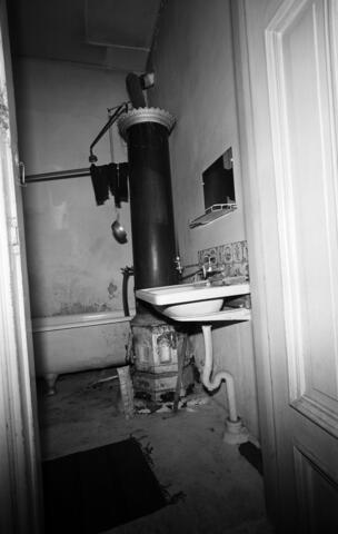 Ett avlångt förfallet badrum med ett badkar längst bak. Framför karet står en pelarlik varmvattenberedare och i förgrunden är en lavoar. 