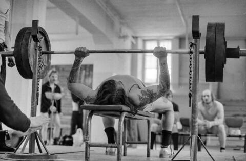 Mies tekee penkkipunnerrusta voimailusalissa. Molemmilla puolilla nostajaa seisovat painojen tukitelineet. Taustalla häämöttää muita nostajia seuraamassa suoritusta ja odottamassa vuoroaan.