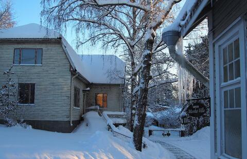 Talvinen piha, jossa näkyy mansardikattoinen, valkoinen puurakennus sekä toisen puurakennuksen kulmaa, jossa räystäästä roikkuu jääpuikkoja. Maassa ja puissa on lunta.