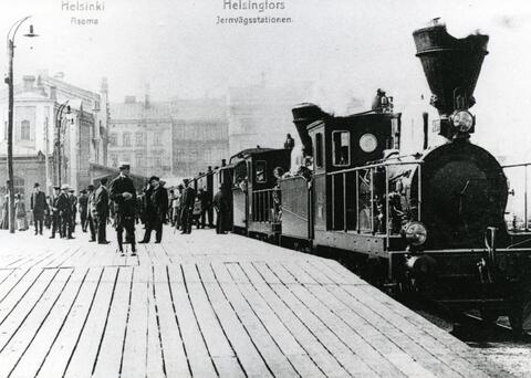 A-sarjan höyryveturit vaunuineen Helsingin vanhalla rautatieasemalla. Asemalaiturilla paljon matkustajia. Helsingin ensimmäisen asemarakennuksen suunnitteli Carl Albert Edelfelt ja se otettiin käyttöön 1862.