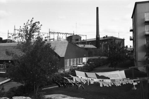 Etualalla pyykkiä kuivumassa narulla kerrostalon pihalla. Taustalla voimalaitoksen rakennuksia, mm. kantaverkon muuntoasema.
