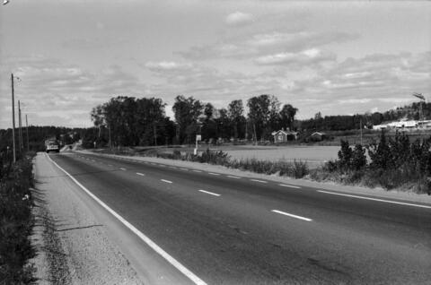 Etualalla on maantie, taaempana peltoa. Keskellä punainen puurakenteinen asuinrakennus Viikin koetilan työntekijöitä varten. Oikella rakenteilla olevaa Helsingin yliopiston Viikin maataloustieteellisten laitosten aluetta. Taaempana Pihlajamäen kallioita.