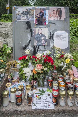Ett kabelskåp med bilder och texter på. Framför skåpet finns det massor av blommor, gravljus, brevlappar, ölburkar och andra minnesgåvor. 
