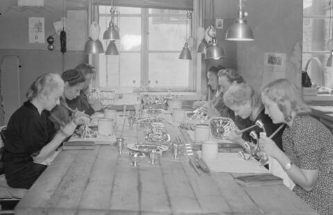 Sju kvinnor bearbetar silverföremål sittande runt ett långt bord vid ett fönster
