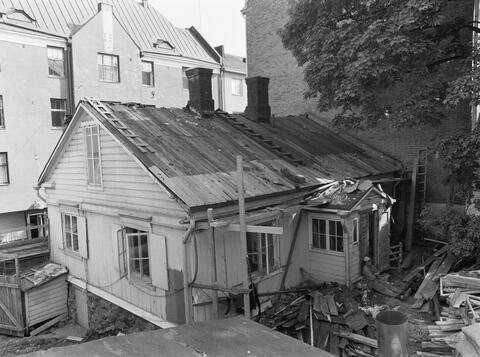 Vanhoja hapertuneita kattohuopakerroksia on poistettu ja osa aluslaudoitusta uusittu. Talon seinustalle on rakennettu työtelineitä. Piha on täynnä rakennusjätettä ja -tarvikkeita. Väsynyt rakentaja istuu portailla nojaten selkäänsä ulko-oveen.