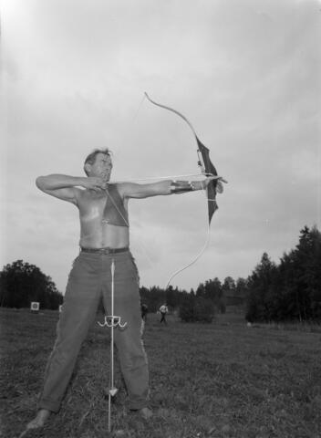Etualalla seisoo mies ilman paitaa virittämässä nuolta jousipyssyyn. Taustalla näkyy maalitauluja ja muita ihmisiä.