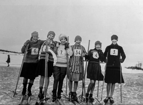 Kuusi hymyilevää naista, joilla on puusukset ja bambusauvat. Jokaisella on rintaan kiinnitetty numerolappu. Yhdellä naisella on pussihousut, muilla on noin polviin asti ulottuva hame. 