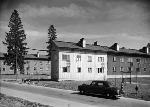 Kaksikerroksisia puutaloja. Etualalla tie, jonka laidassa on pysäköity henkilöauto. Lapsia leikkii ojissa ja pihoilla.