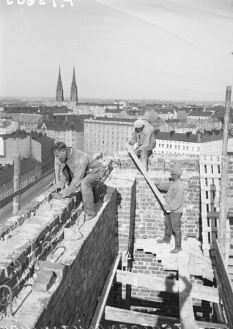 Kolme miestä työskentelee korkealla kirkonrakennustyömaalla. Taustalla näkyy kerrostalojen yläkerroksia ja kattoja sekä Johanneksenkirkon kaksi tornia.