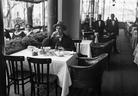 En man sitter vid ett dukat bord och dricker. Bakom ett räcke till vänster sitter två kvinnor. I bakgrunden syns två servitörer.