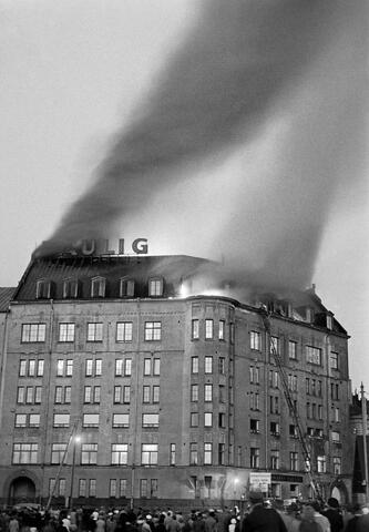 A building with a burning attic. A lot of people have gathered on the street to watch it.