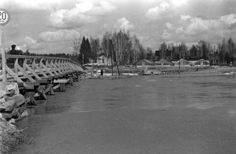 Vasemmalla on puinen silta, oikealla etualalla tulviva Vantaanjoki. Maantiellä ajaa linja-auto, taustalla on kasvihuoneita ja puisia huviloita.