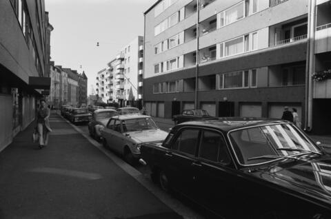 Gatan omges av nya flervåningshus. Lägs gatan är flera bilar parkerade och några fotgängare går på trottoarerna.