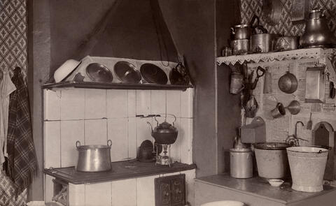 Vasemmalla on puuhella, oikealla on senkki, jonka päällä on sankoja. Seinällä on keittiövälineitä ja seinällä kattilahylly.