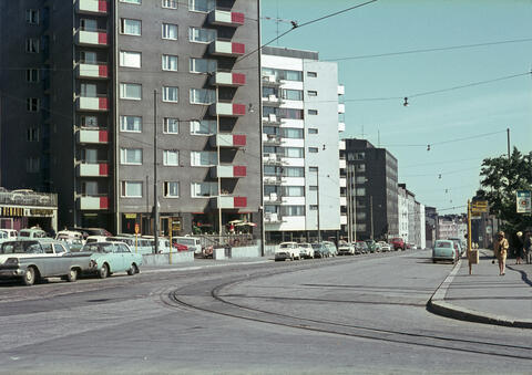 På färgfotografiet, som är taget på sommaren, syns höghus i nio våningar. På gatan finns bilar och fotgängare.