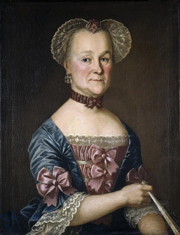 A half-body portrait of a 59-year-old woman in formal attire. A blue dress, an open-fronted coat-like garment covering the shoulders, with cold red silk roses and light lace on the sleeves and at the neckline. On her head, the woman is wearing a lace-edged red butterfly cap, with a brown silk bow around her neck and a light-coloured fan in her hand.