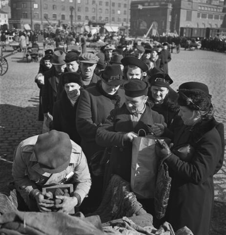 En lång kö av främst kvinnor på Hagnäs torg väntar på sin tur för potatisdistribution. I förgrunden en man i keps som mäter potatis med en träkappa